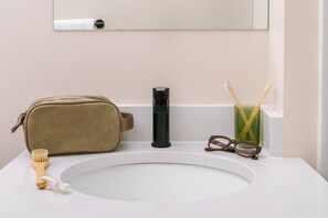 Un lavabo de salle de bain avec un sac beige, une bouteille noire, une brosse à dents et des lunettes posés sur le comptoir.