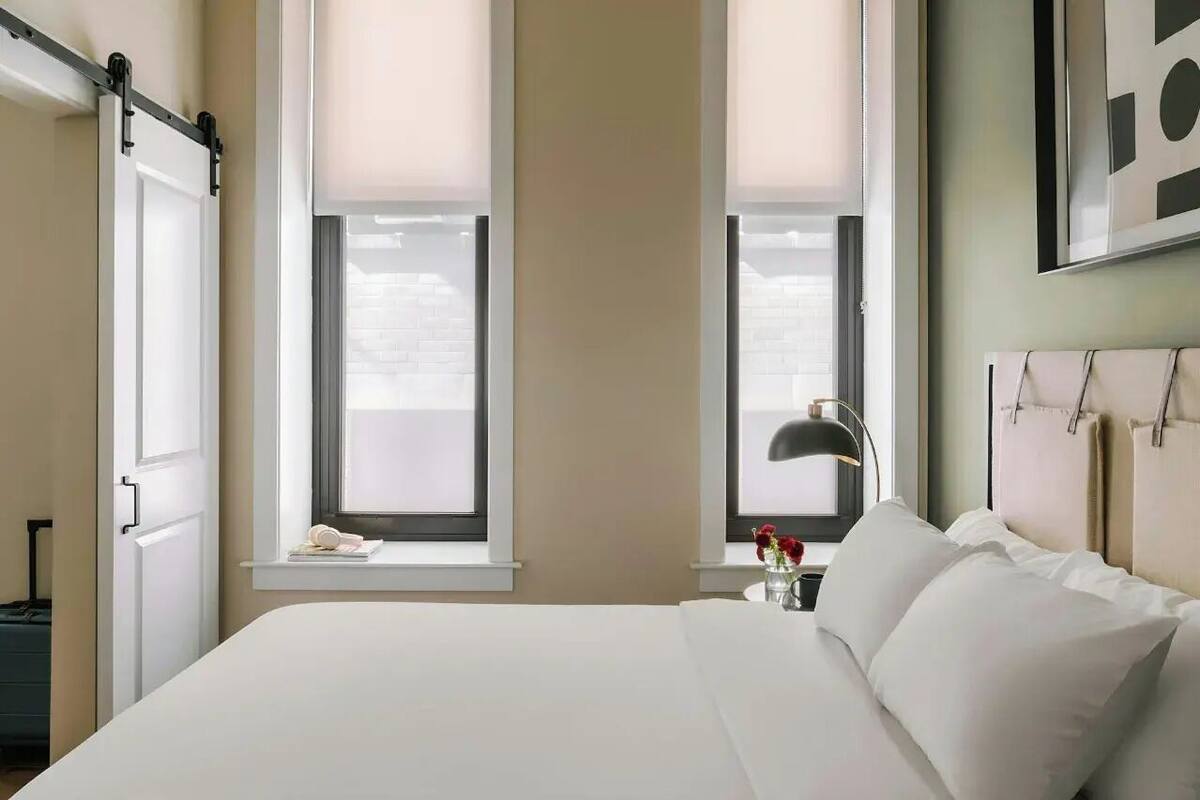 Ett hotellrum med en säng, två fönster med persienner, ett sängbord med en lampa och ett konstverk som hänger på väggen.