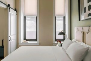 Habitación de hotel con cama, dos ventanas con persianas, una mesita de noche con lámpara y una obra de arte colgada en la pared.