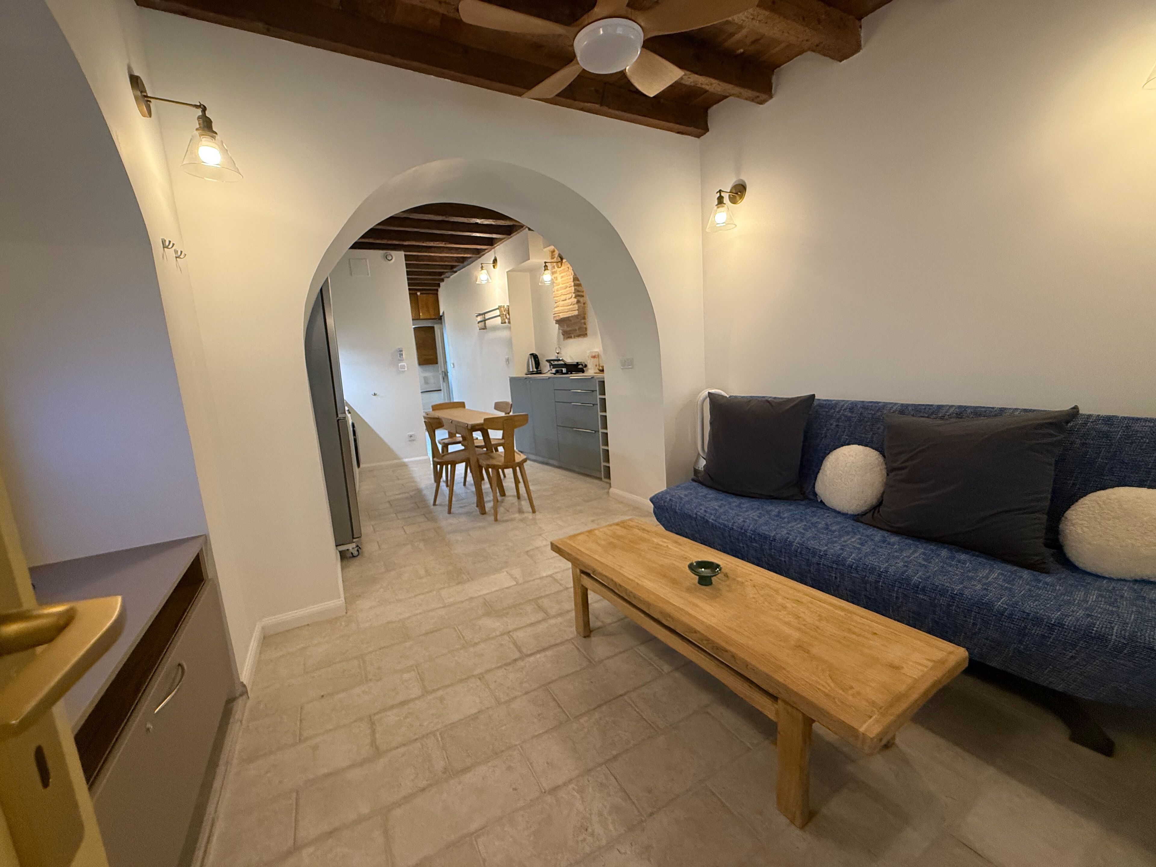 A cozy living area with a wooden bench, a blue sofa with cushions, and an arched doorway leading to a kitchen.