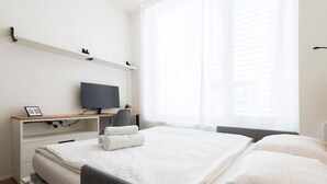A hotel room with a bed, a desk with a computer, a chair, and a window with blinds. - SMART studio w/1GB speed WiFi + Monitor | A/C | Onsite Free Parking Spot (Praha 5)