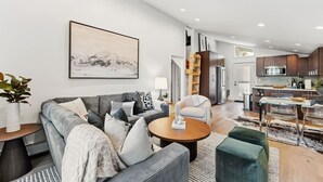 Un salon moderne avec un canapé gris, une table basse et un coin repas comprenant une table et des chaises en verre. On aperçoit, au fond, une cuisine avec des meubles sombres et des appareils électroménagers en acier inoxydable.