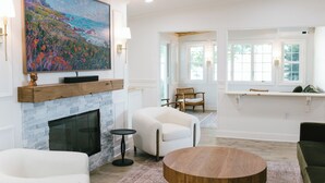 A spacious living room with a fireplace, a large painting, and a view of the outdoors through a window.