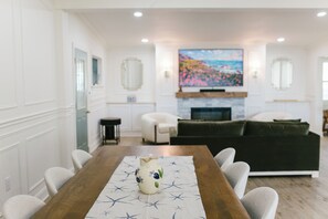 A dining area with a wooden table, chairs, and a vase centerpiece. A living room with a sofa and a painting is visible in the background.