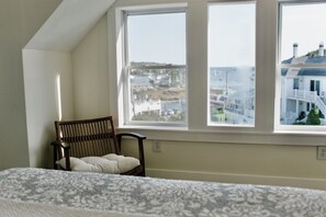 A bedroom with a large window overlooking a coastal town, a wooden chair, and a patterned bedspread.