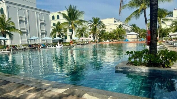 Piscina com espreguiçadeiras e guarda-sóis, cercada por palmeiras e um edifício com janelas em arco.