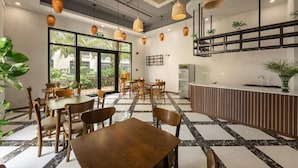 Um café moderno com mesas e cadeiras de madeira, chão em piso preto e branco, luminárias de vime pendentes e um balcão com uma planta.