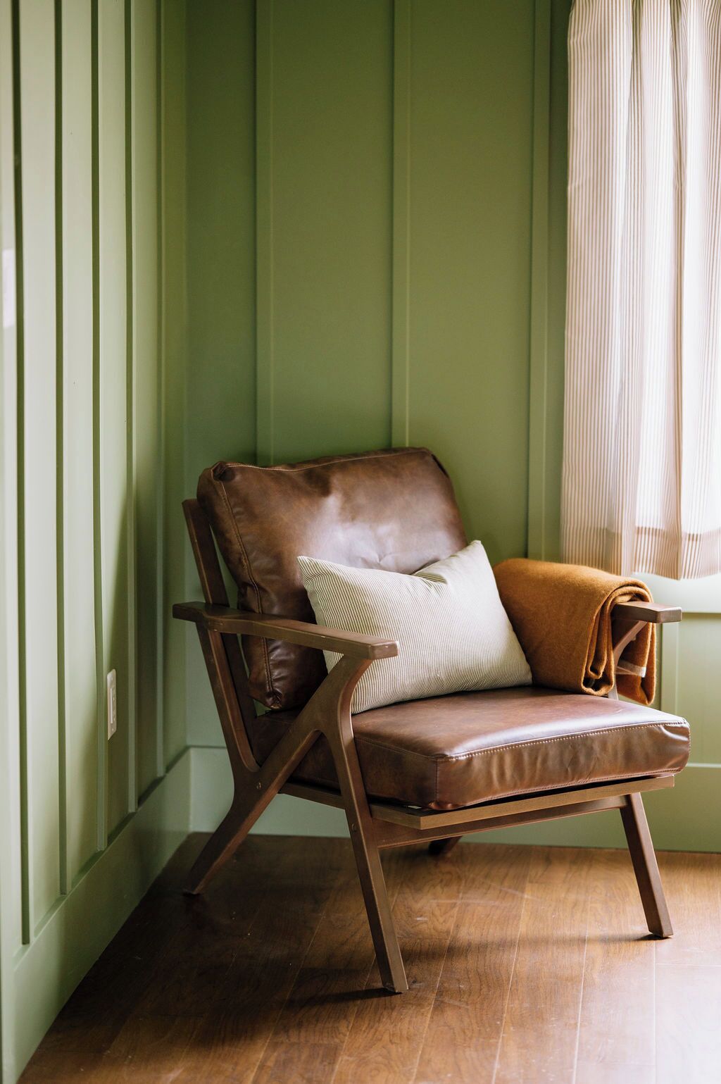 A brown leather chair with a cushion and a blanket, positioned against a green wall with a window.
