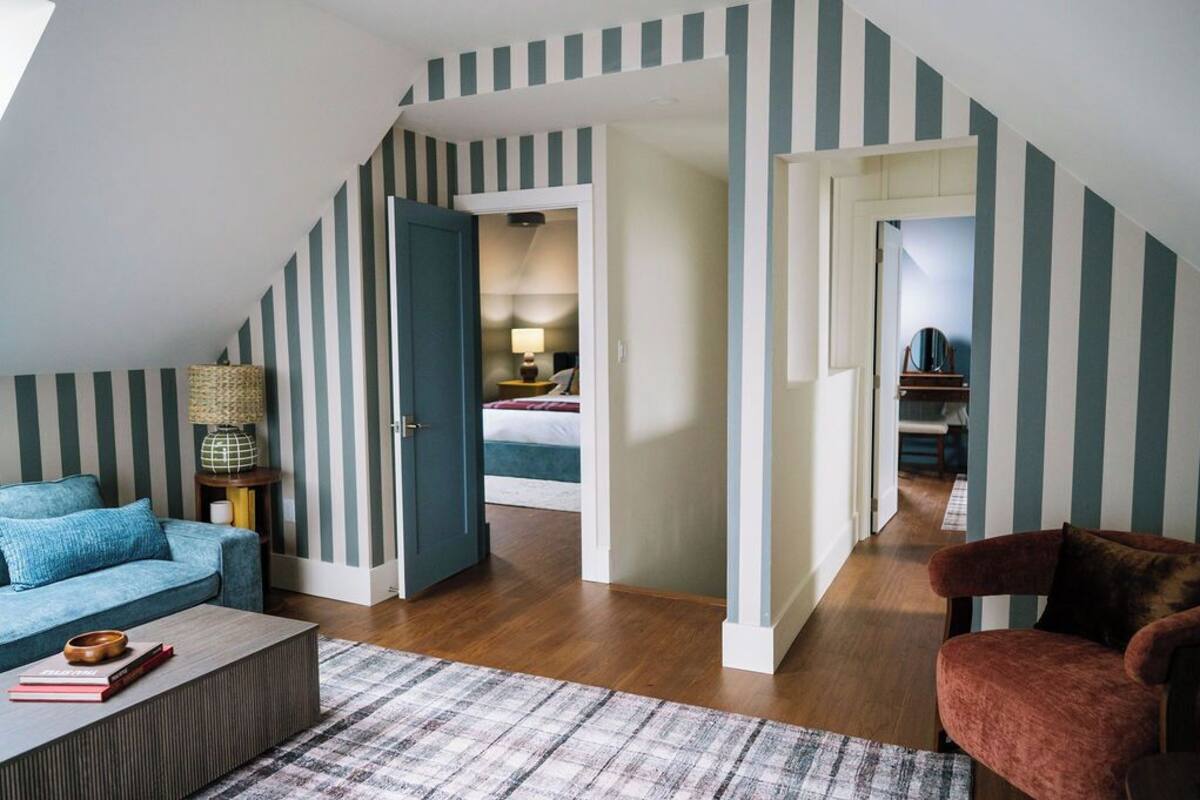 Un dormitorio estilo loft con techo inclinado, paredes a rayas, un sofá azul, un sillón rojo y una zona de dormitorio visible con cama y mesita de noche.