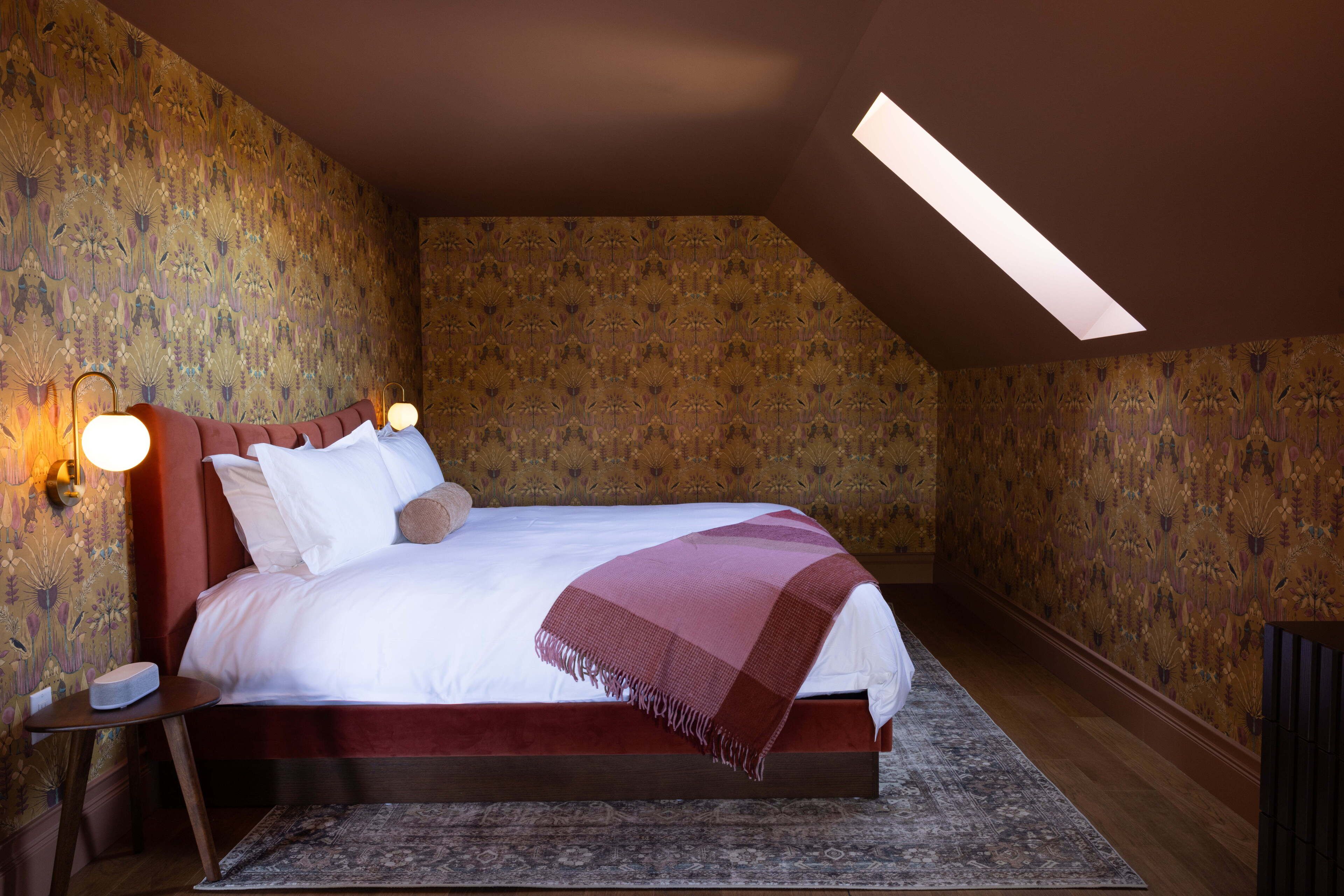 A bedroom with a large bed, a skylight, patterned wallpaper, and a nightstand with a lamp.