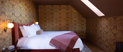 A bedroom with a large bed, a skylight, patterned wallpaper, and a nightstand with a lamp.
