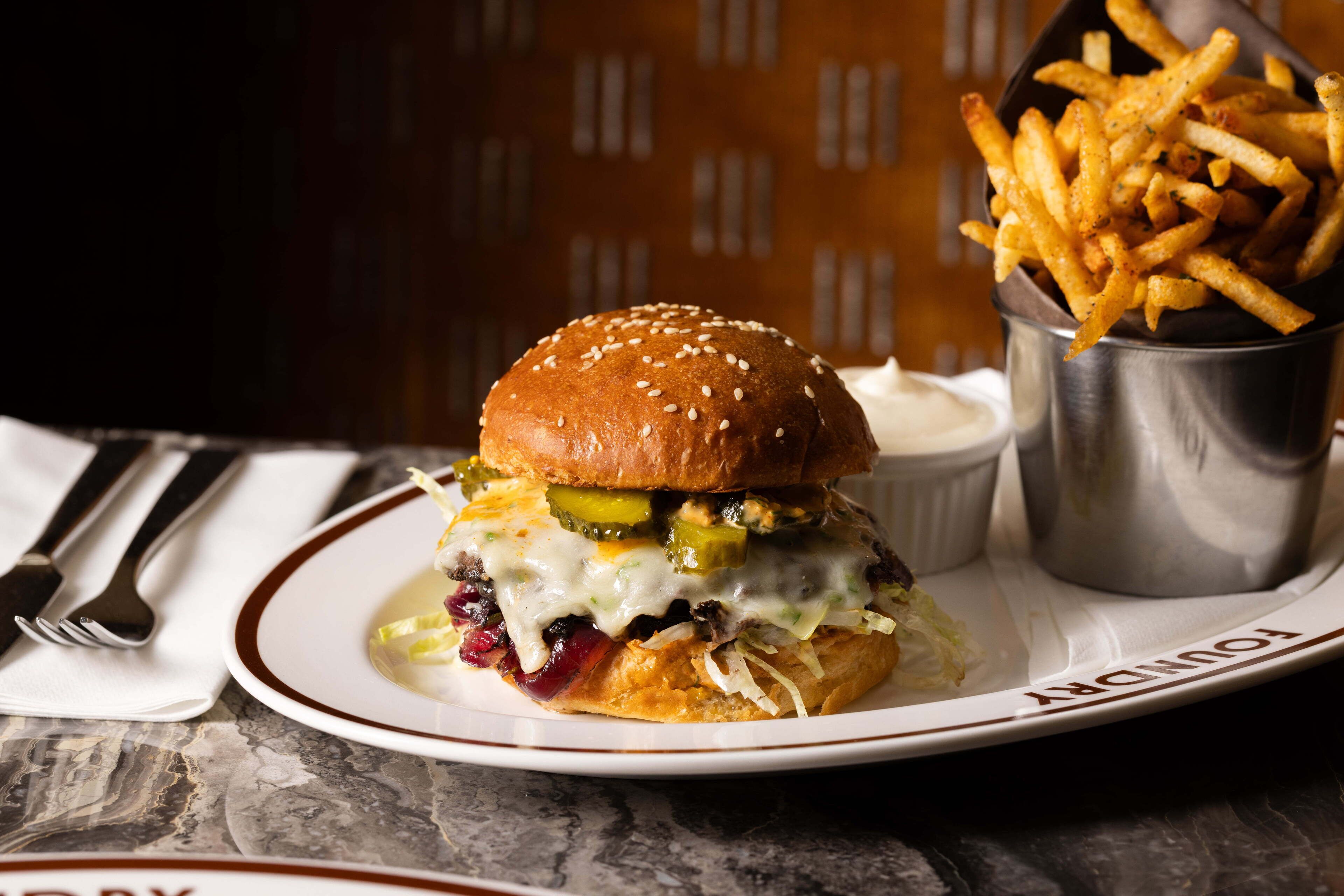 A burger with cheese, pickles, and a bun on a plate, accompanied by a side of fries and a small container of sauce.