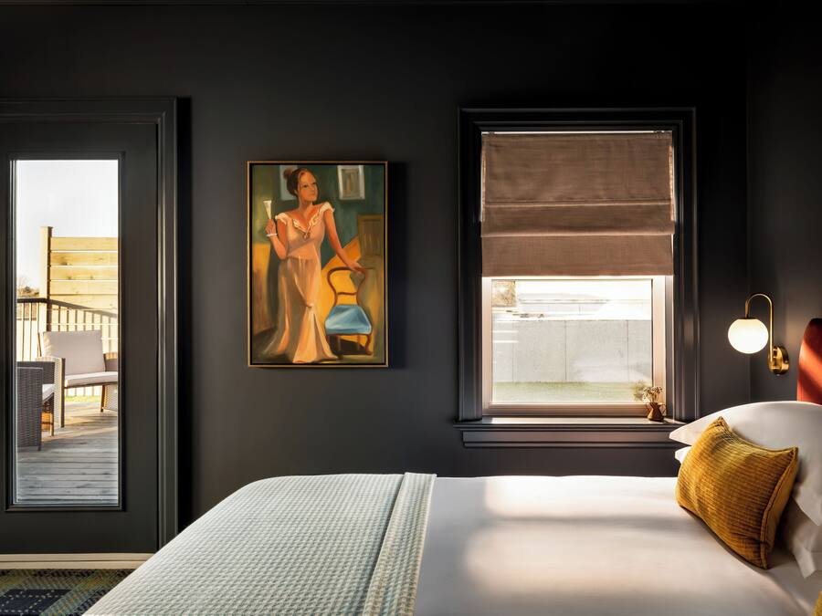 A hotel room with a bed, a painting, a window with a view, and a door leading to an outdoor area.