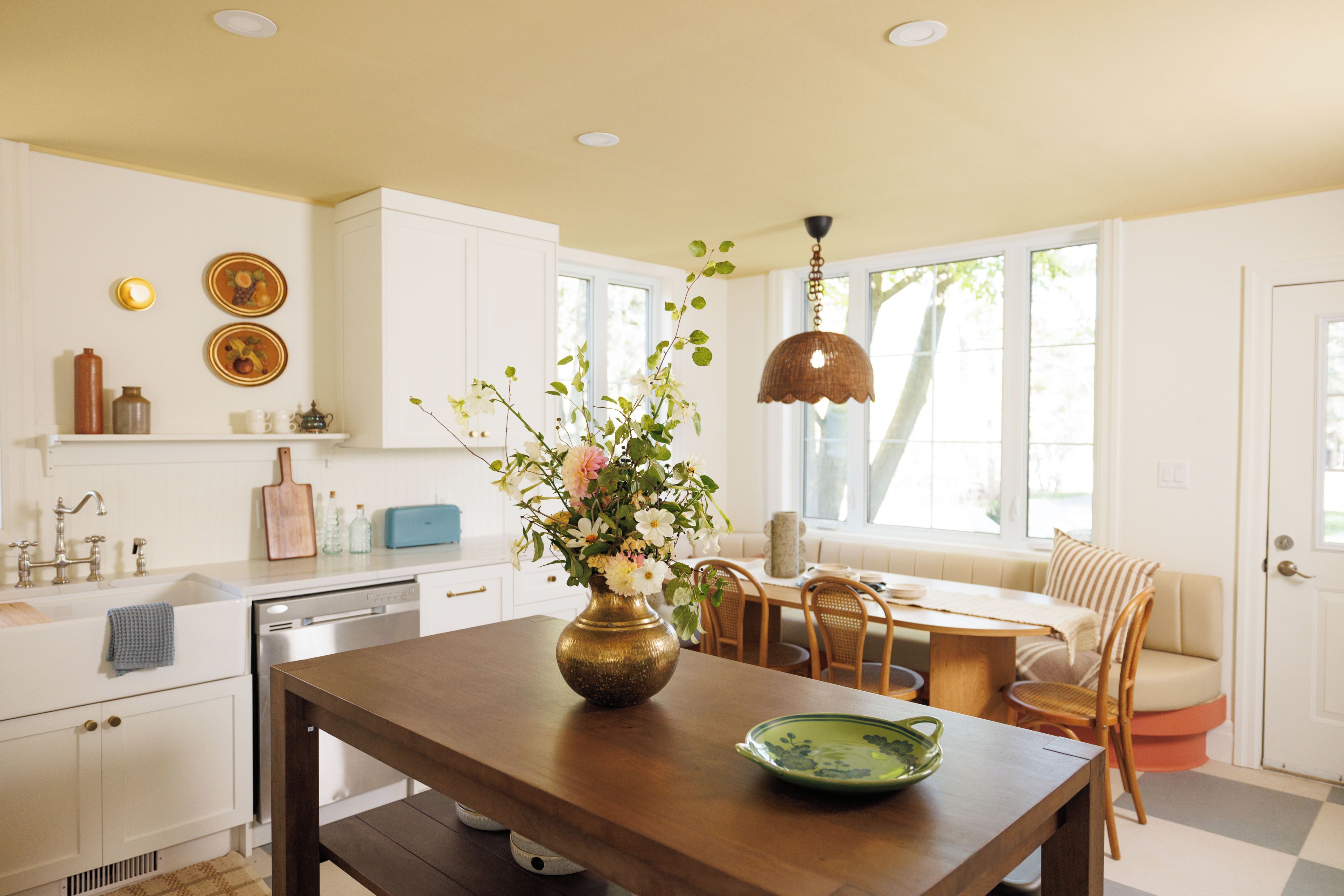A kitchen with white cabinets, a wooden dining table, a vase with flowers, and a hanging light fixture.