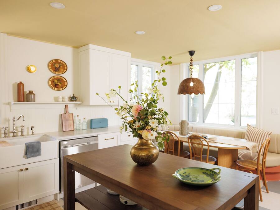 A kitchen with white cabinets, a wooden dining table, a vase with flowers, and a hanging light fixture.