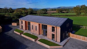 A modern two-story building with a sloped roof, surrounded by a gravel driveway and grassy areas. - The Gathering Barn | Hot Tub | Sleeps 12 | Luxury Countryside Views | (Hereford)