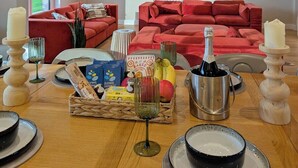 A spacious living room with a red sofa set, a dining table with candles, a bottle of champagne, and a basket of bread. - The Gathering Barn | Hot Tub | Sleeps 12 | Luxury Countryside Views | (Hereford)
