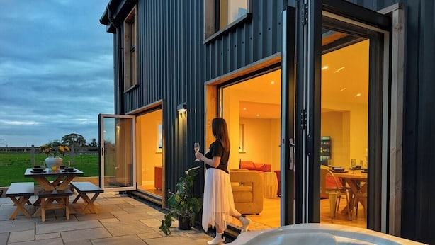 A modern house with a hot tub, a dining area, and a person walking towards the entrance. - The Gathering Barn | Hot Tub | Sleeps 12 | Luxury Countryside Views | (Hereford)