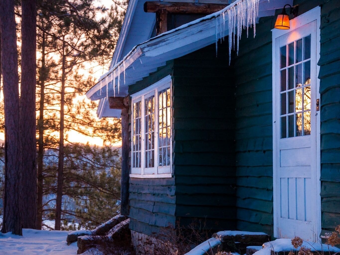 A wooden cabin with a snow-covered porch, a window with a white frame, and icicles hanging from the roof.