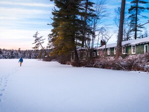 A snow-covered landscape with a person walking away from the camera, a green building with white trim, and a forest in the background. - White Pine Camp (Rainbow Lake)