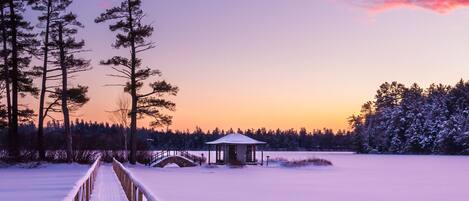 A snow-covered path leading to a small wooden structure with a roof, surrounded by tall trees and a frozen lake.