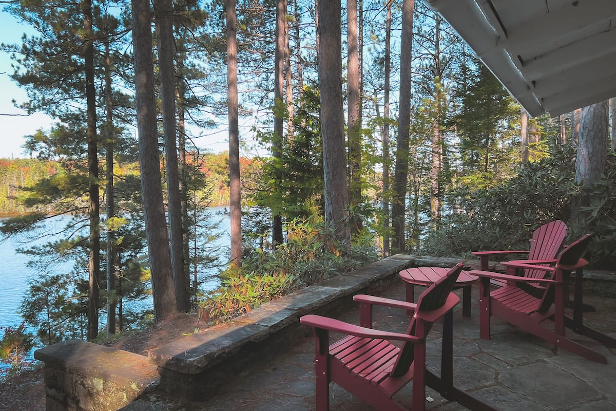 Una terraza de madera con sillas Adirondack con vistas a un lago rodeado de altos árboles.