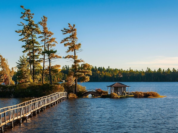 A wooden pier extending into a lake, surrounded by tall trees and a small gazebo on an island. - White Pine Camp (Rainbow Lake)