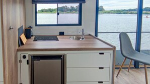 Une cuisine compacte équipée d’une cuisinière intégrée, d’un évier et d’un petit réfrigérateur. Elle dispose d’une grande fenêtre offrant une vue sur un lac et des arbres.