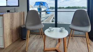 Une pièce moderne avec un plancher en bois, deux chaises grises, une petite table blanche et une fenêtre donnant sur un bateau et l’eau.