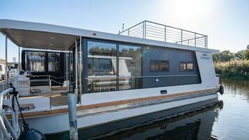 Ein modernes Hausboot mit überdachter Terrasse, großen Fenstern und einem Metallgeländer.