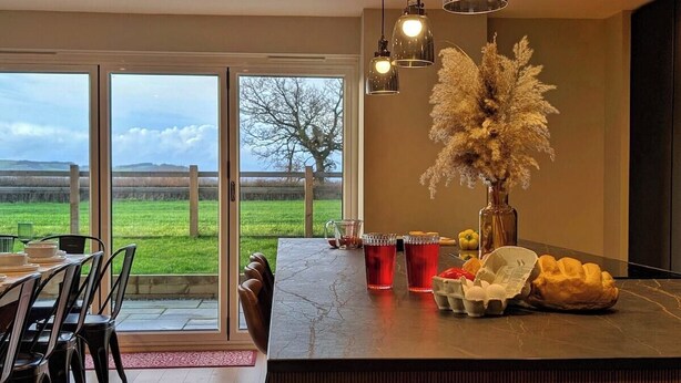 Una cocina moderna con una isla grande, un área de comedor con sillas y vistas a un paisaje verde a través de amplios ventanales.