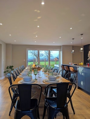 A dining area with a long table set for a meal, surrounded by chairs, with a view of a green landscape through large windows.