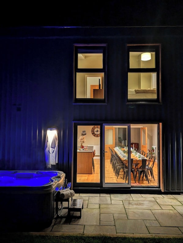 A modern house with a hot tub, a dining area with a large table and chairs, and a kitchen visible through the glass doors.