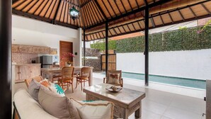 Un vaste espace de vie comprenant un grand canapé, une table à manger et des chaises. On y trouve également une cuisine équipée de placards et un téléviseur. Une piscine est visible au fond, entourée d’un mur blanc et de verdure.