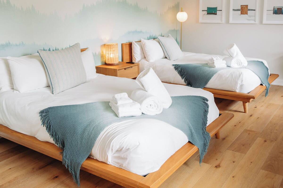 Habitación de hotel con dos camas, suelo de madera, un mural temático de montañas, lámparas de noche y cuadros en la pared.