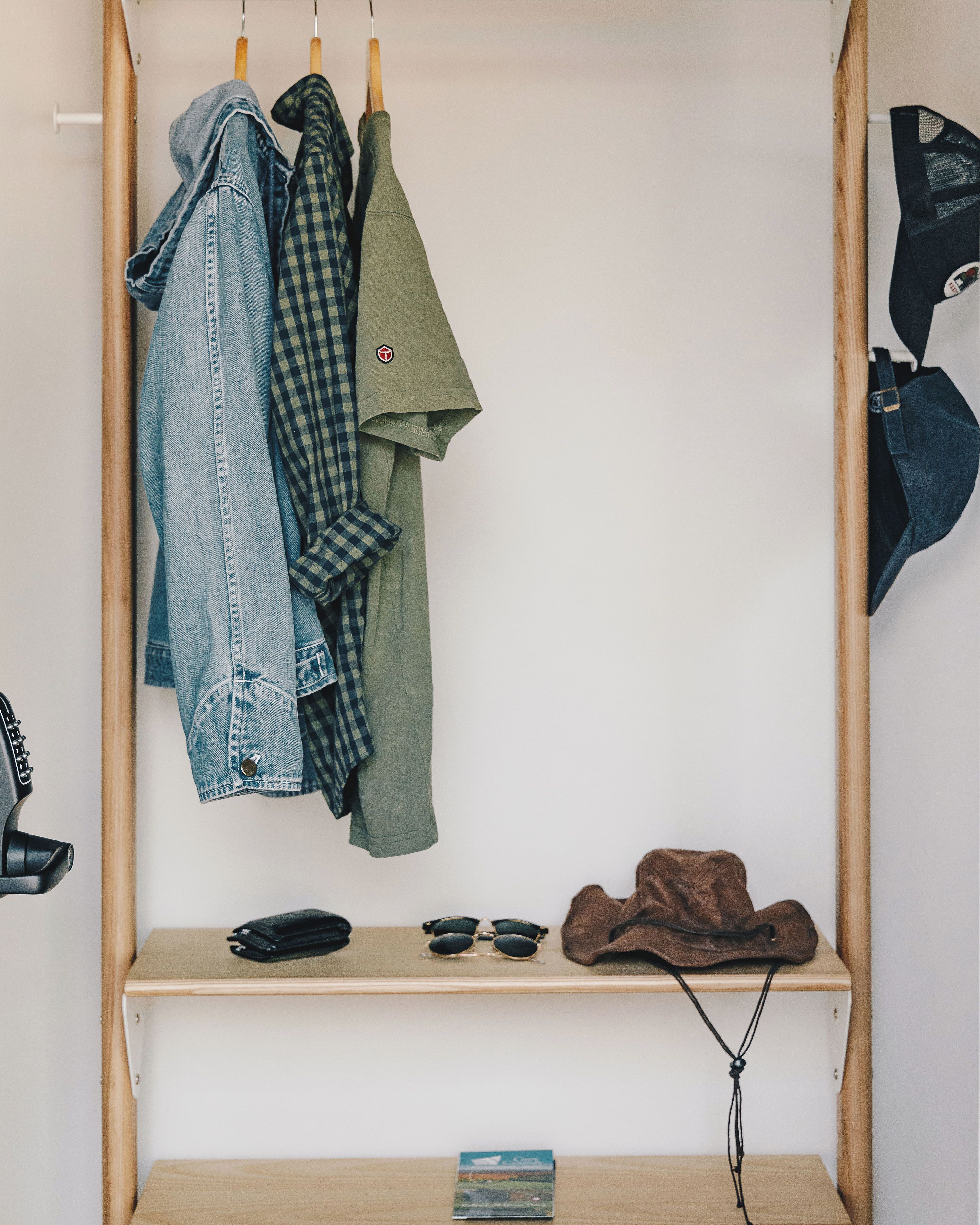 A wooden wardrobe with hanging clothes and a shelf with sunglasses, a hat, and a wallet.
