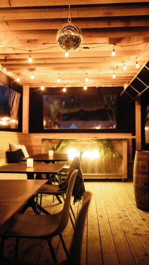 Eine Holzdachterrasse mit Tischen und Stühlen, einer hängenden Discokugel und einem großen Bildschirm, der eine Nachtszene zeigt.