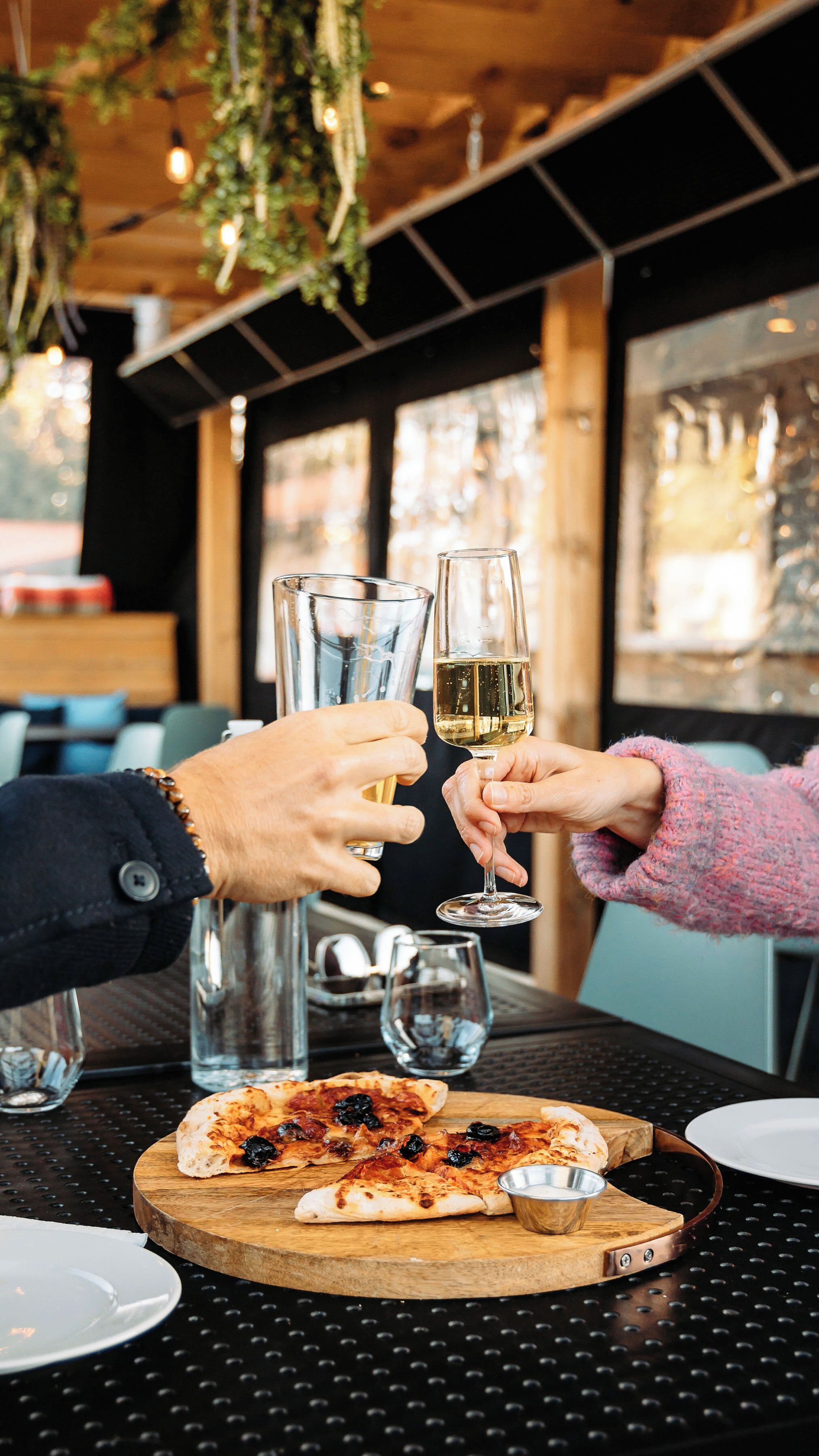 Un ambiente de restaurante con una mesa de madera, una pizza y dos personas brindando con vasos.