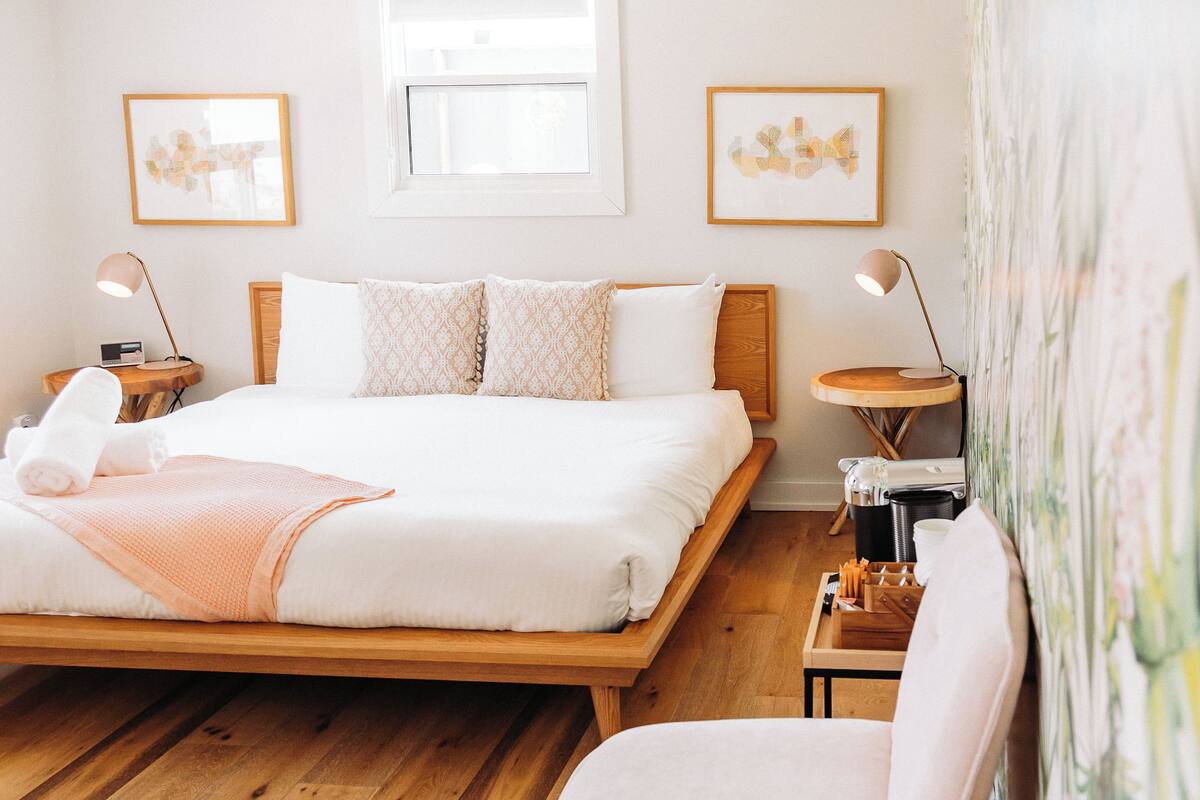 Una habitación de hotel ordenada con cama, mesitas de noche, una silla, un taburete de madera redondo y obras de arte en las paredes.