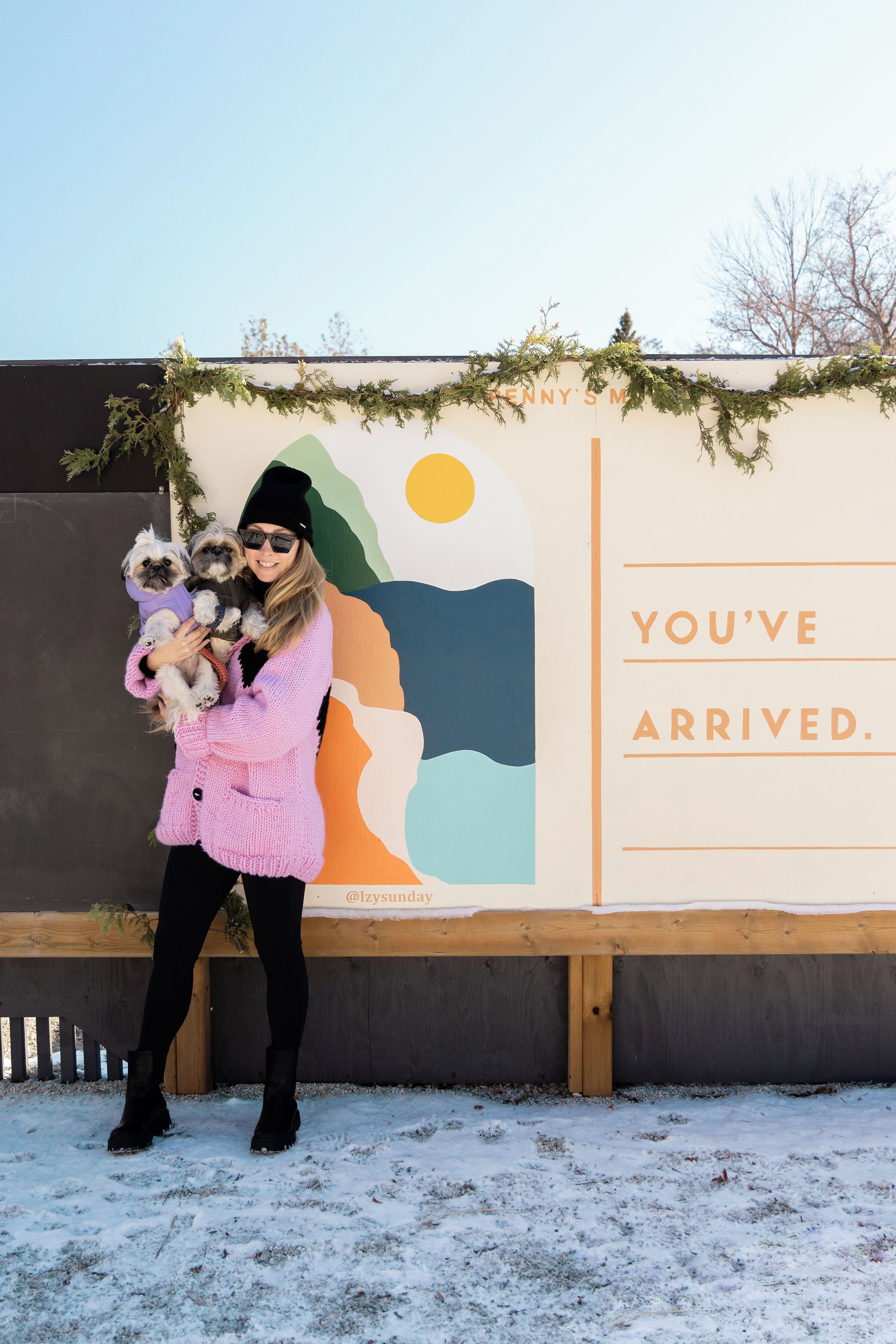 A person holding two dogs in front of a wall with a sign that reads "You've arrived."