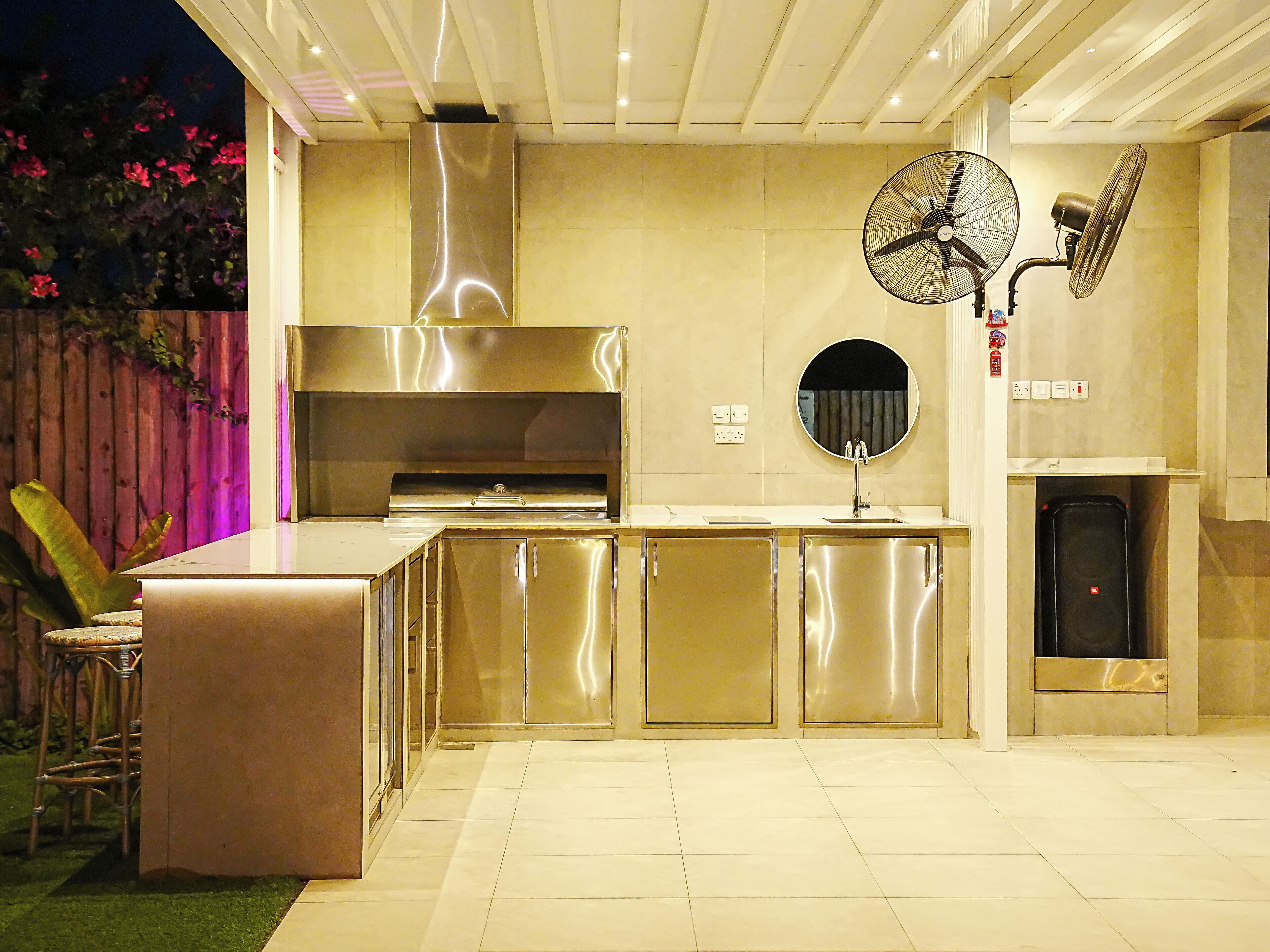 A modern outdoor kitchen with a stainless steel range hood, a built-in sink, and a round mirror above it.