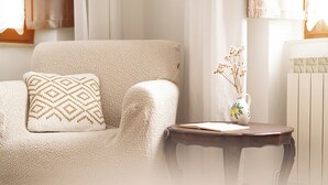 Un sillón color beige con un cojín estampado, una mesita de madera con un jarrón de flores secas y un radiador al fondo.