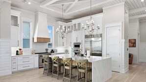 Una cucina moderna con mobili bianchi, un'isola grande con sgabelli da bar, elettrodomestici in acciaio inossidabile e un lampadario.