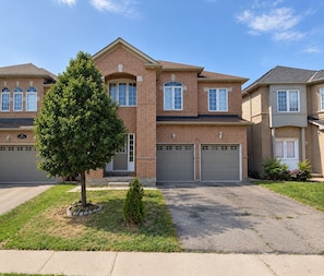 A two-story brick house with a garage, a small tree in the front yard, and a clear sky. - Private Queen Bedroom w/ Walk-In Closet, TV & Mini Fridge, 2.5 Bath—Shared Home (Brampton)