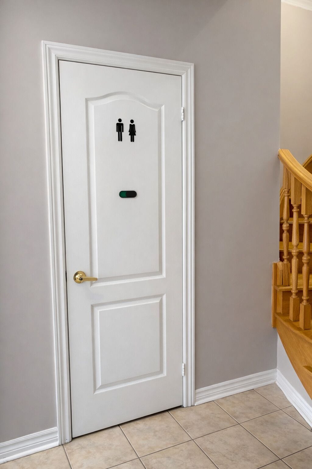 Una puerta blanca con un símbolo de género y un botón verde, una escalera de madera y suelo con baldosas color beige.
