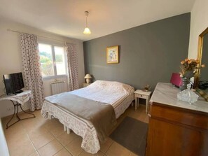 Ein Schlafzimmer mit Bett, Schreibtisch mit Computer, Kommode und Fenster mit Vorhängen.