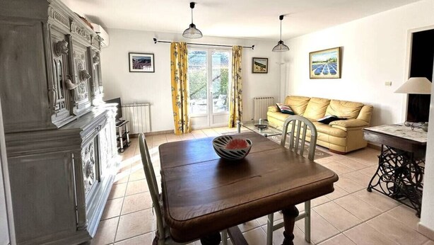 Un comedor con una mesa y sillas de madera, un jarrón con flores secas, un radiador, un sofá y un cuadro en la pared.