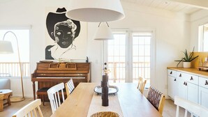 Un comedor amplio con una mesa de madera larga, sillas blancas, un piano y una lámpara colgante grande.