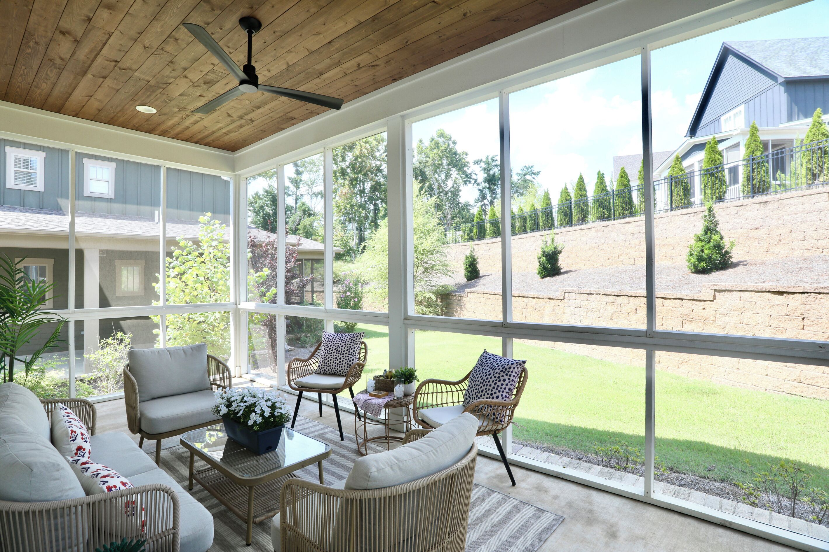 Un patio couvert aménagé avec des meubles en osier, un ventilateur de plafond et de grandes fenêtres donnant sur un jardin paysager.