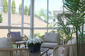 A sunlit room with wicker furniture, a glass table, and a view of a hillside. - "Sippin on Savannah Square" by OVR (Oxford)